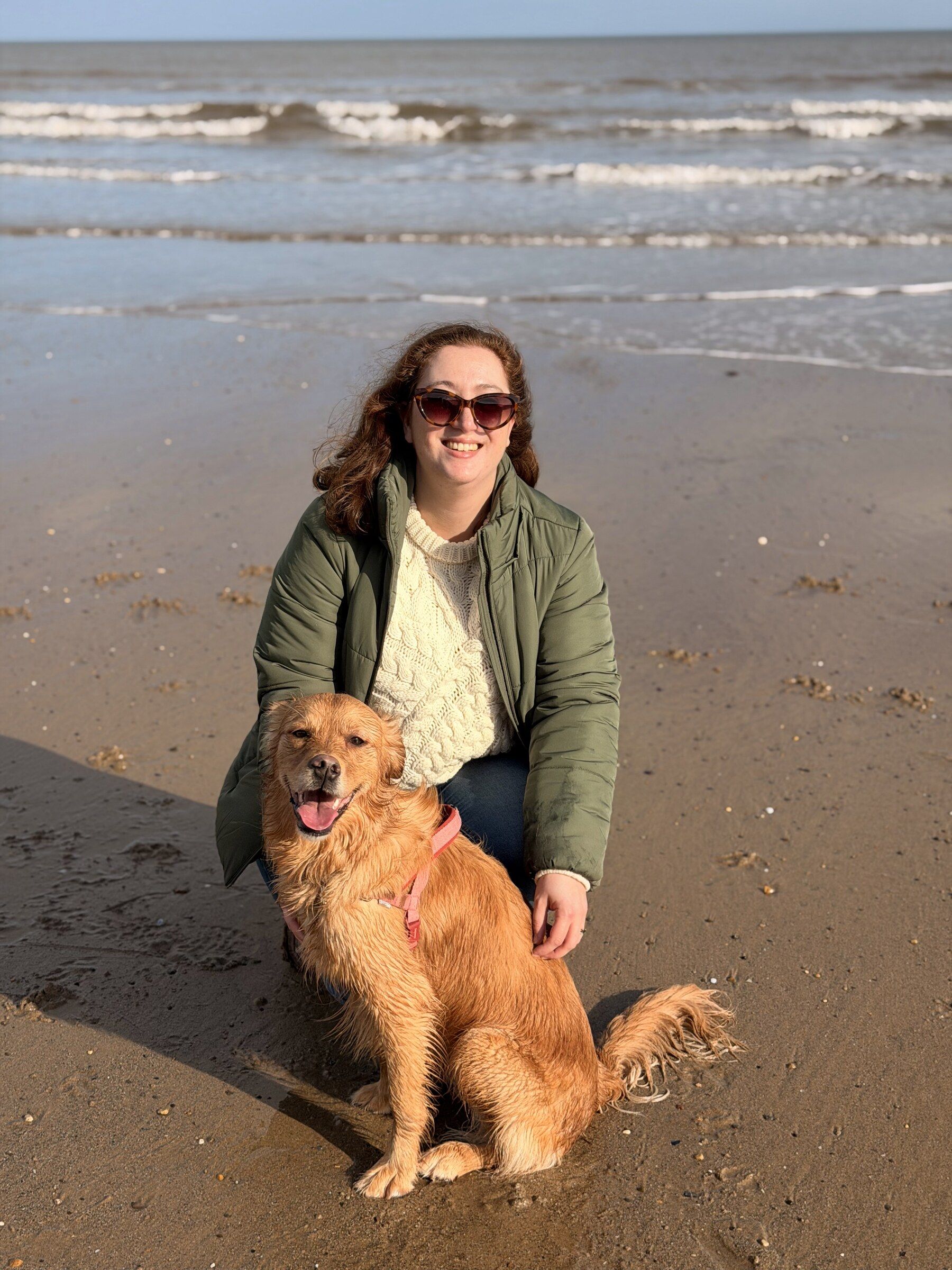Tamara crouching beside a golden retriever on wet sand with the sea behind them